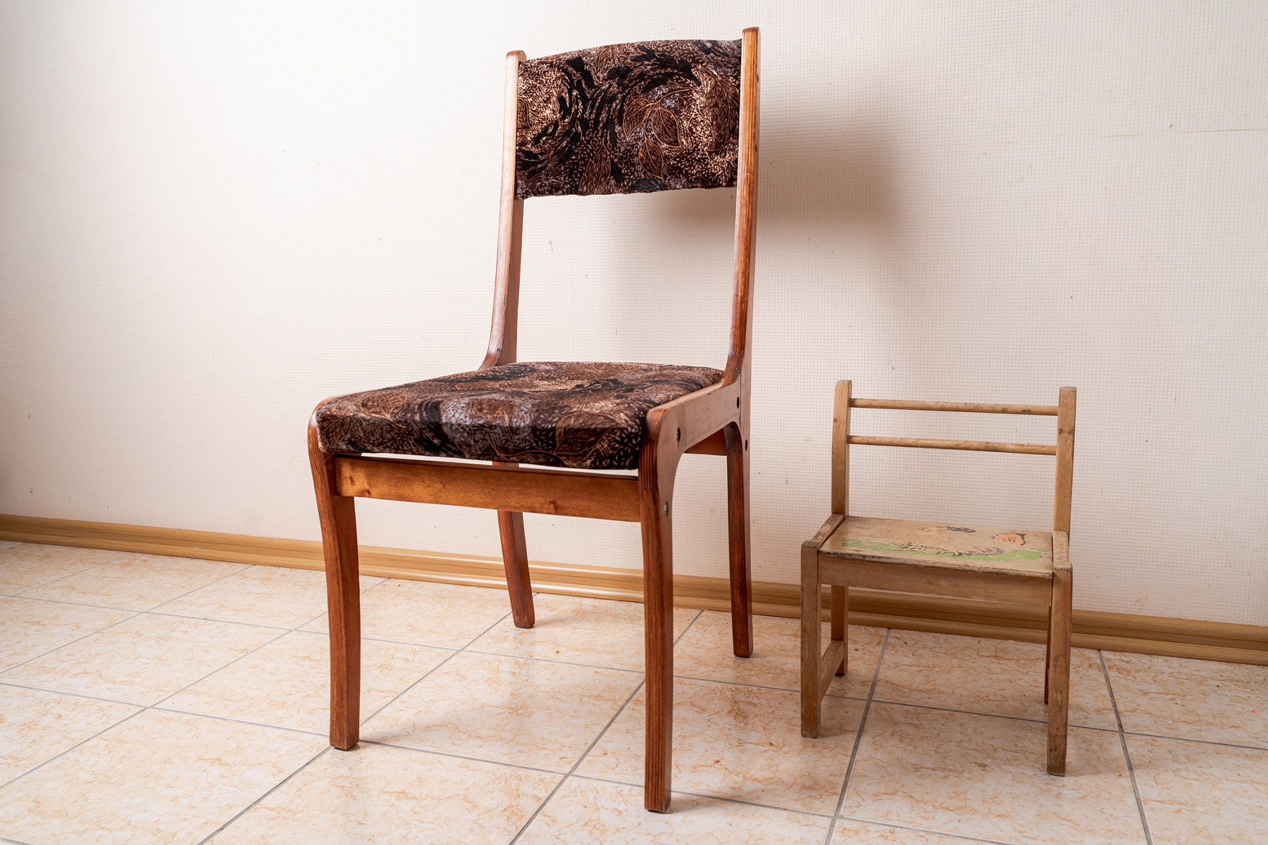 Ein Kinderstuhl und ein großer Stuhl, beide aus Holz, stehen auf einem Fliesenboden. Hintergrund weiße Wand.